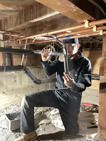 Victor kneeling in a crawlspace fitting a new supply line beneath copper pipes