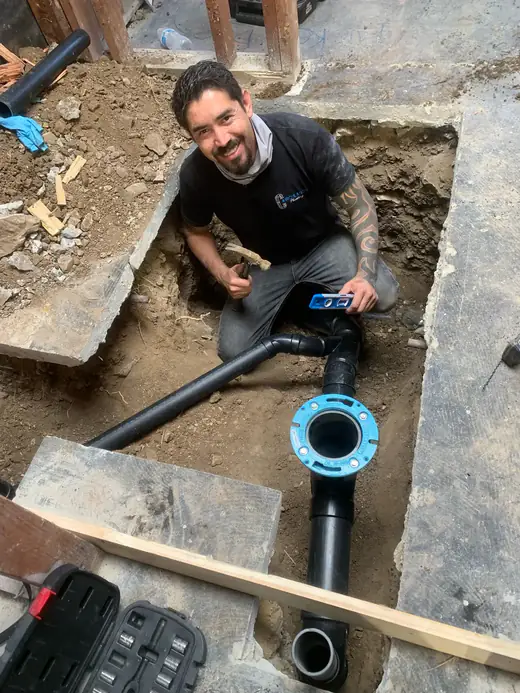 Victor Herrera smiling in an excavated trench beside new black ABS drain pipe and a toilet flange